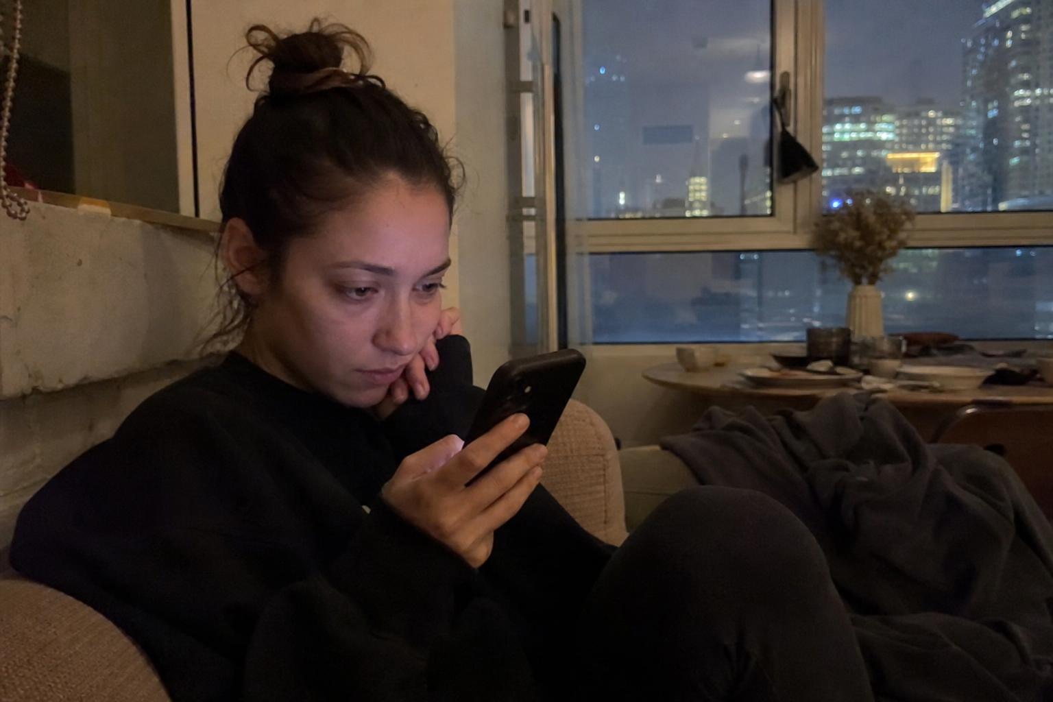 A young woman alone in a dim apartment at night, looking cautiously at her phone with a concerned expression while staying safe during an anonymous online chat