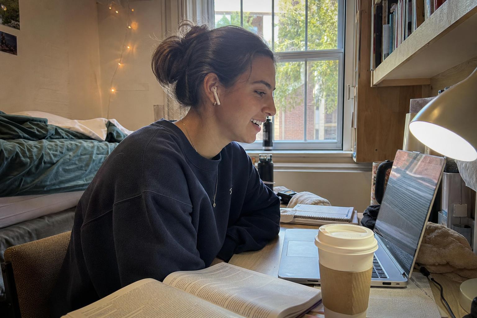 A college student in a dorm room smiling while meeting new people online through a video chat on her laptop during back-to-school season
