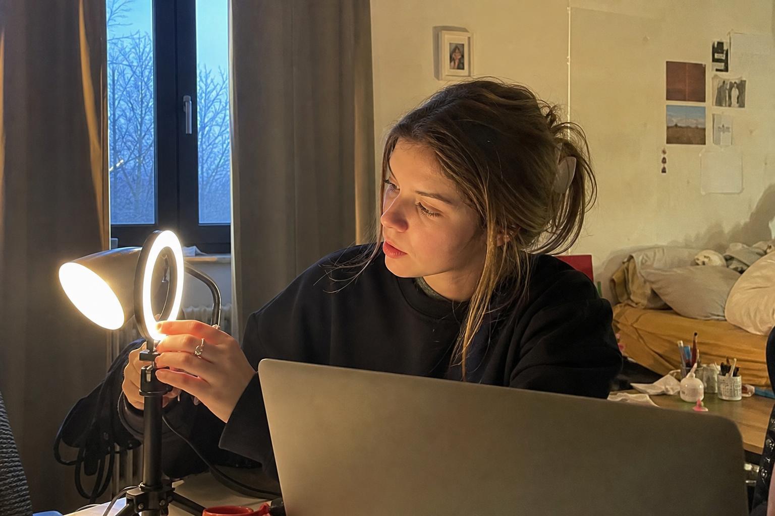 A young woman adjusting a ring light and desk lamp beside her laptop in a cozy dim bedroom, setting up good lighting and a tidy background before a random video chat call