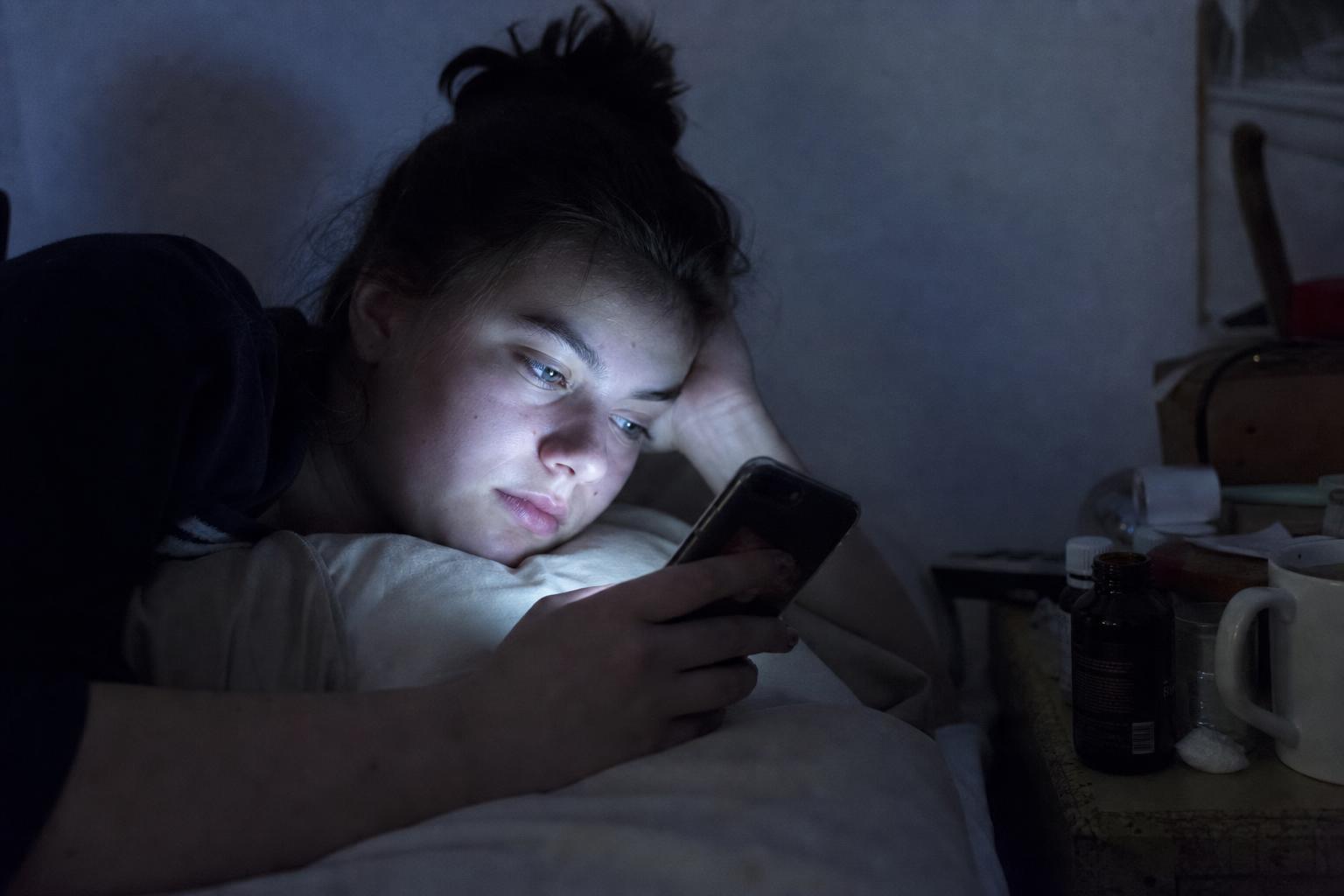 Young woman lying in bed at night in a dimly lit room, face lit by her phone screen during a private anonymous chat