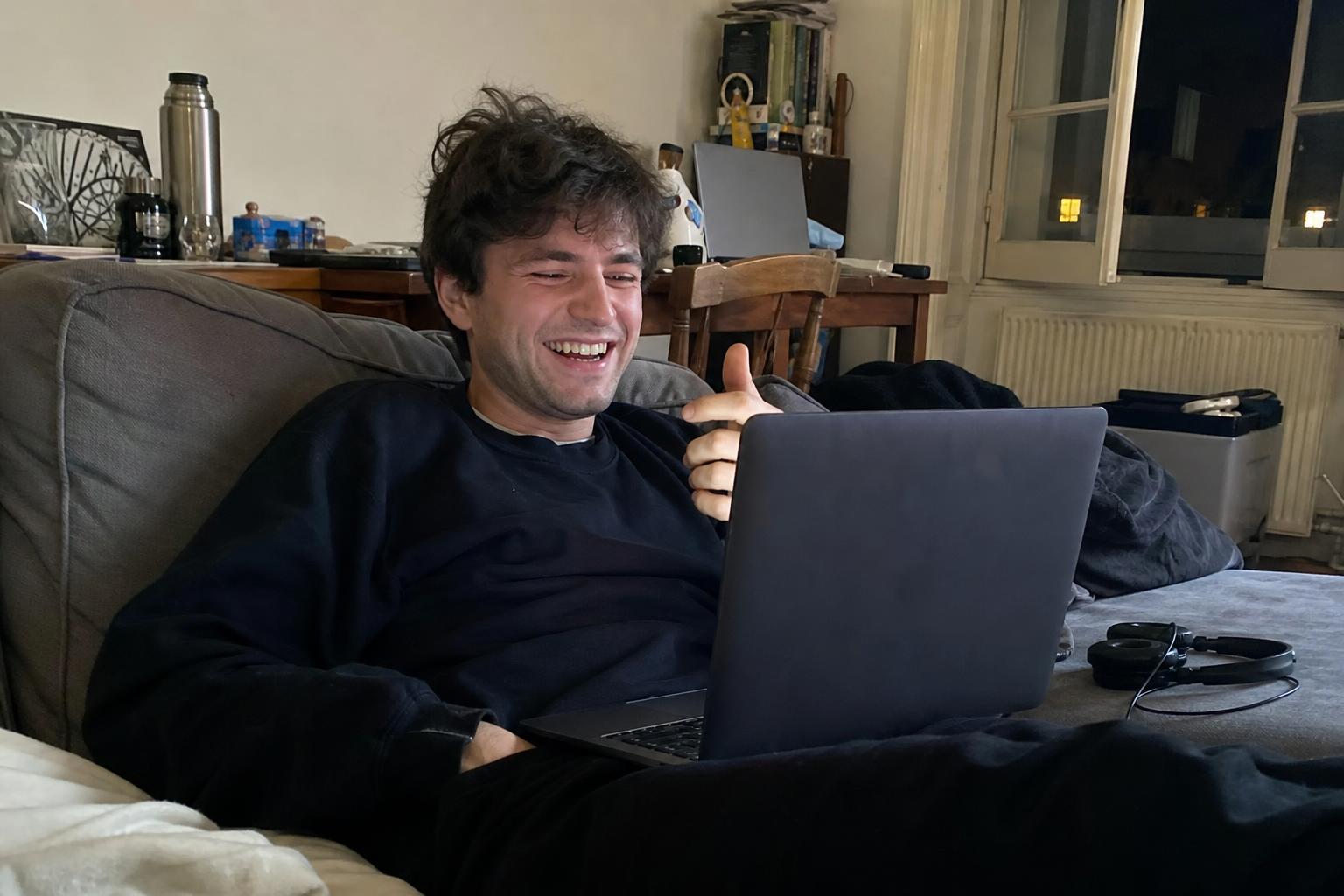 Young man laughing on his couch while he talks to strangers online via video chat on his laptop in the evening